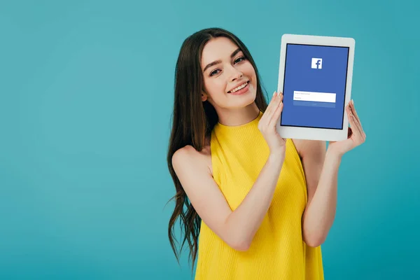 KYIV, UCRANIA - 6 de junio de 2019: hermosa chica feliz en vestido amarillo mostrando tableta digital con aplicación de Facebook aislada en turquesa - foto de stock