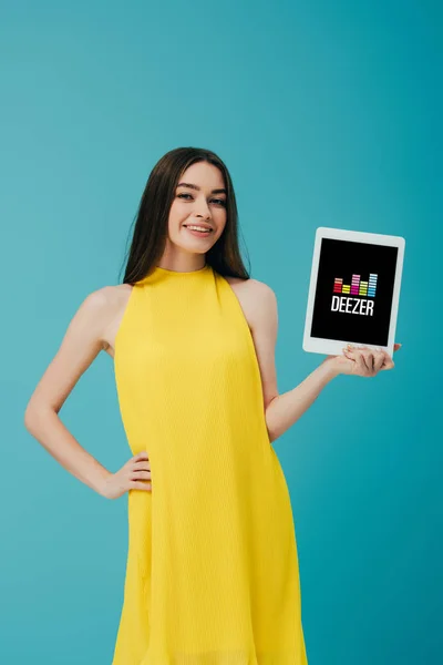 КИЕВ, УКРАИНА - 6 ИЮНЯ 2019 года: улыбающаяся девушка в желтом платье с рукой на бедре показывает цифровой планшет с приложением Deezer изолированы на бирюзовом — стоковое фото