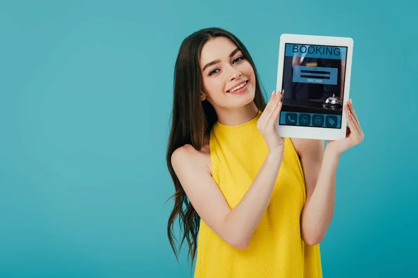 Glückliches brünettes Mädchen im gelben Kleid zeigt digitales Tablet mit Buchungs-App isoliert auf Türkis — Stockfoto