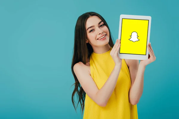 KYIV, UCRANIA - 6 de junio de 2019: hermosa chica feliz en vestido amarillo que muestra la tableta digital con la aplicación snapchat aislada en turquesa - foto de stock