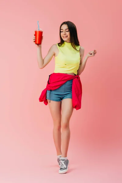 Vista completa de la hermosa chica feliz en ropa brillante sosteniendo soda en taza de plástico aislado en rosa - foto de stock