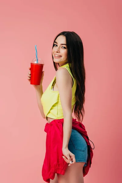 Hermosa chica feliz en ropa brillante sosteniendo soda en taza de plástico aislado en rosa - foto de stock