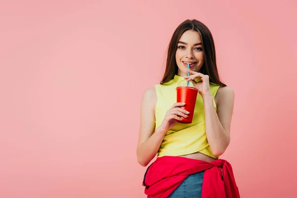 Beautiful smiling girl in bright clothes drinking soda from straw isolated on pink — Stock Photo