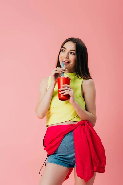 Beautiful smiling girl in bright clothes drinking soda from straw isolated on pink — Stock Photo