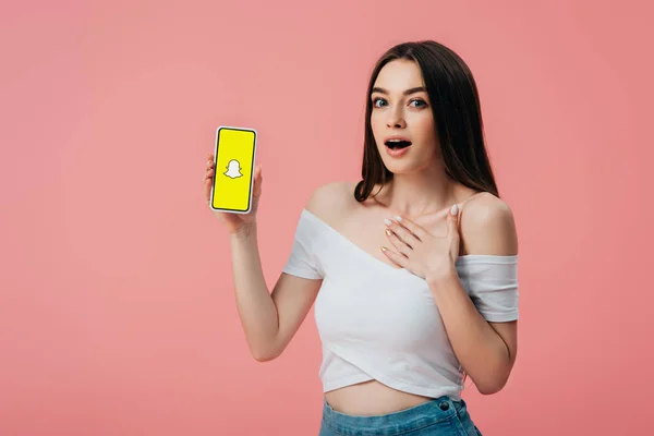 KYIV, UCRAINA - 6 GIUGNO 2019: bellissima ragazza scioccata che tiene lo smartphone con l'app snapchat isolata su rosa — Foto stock