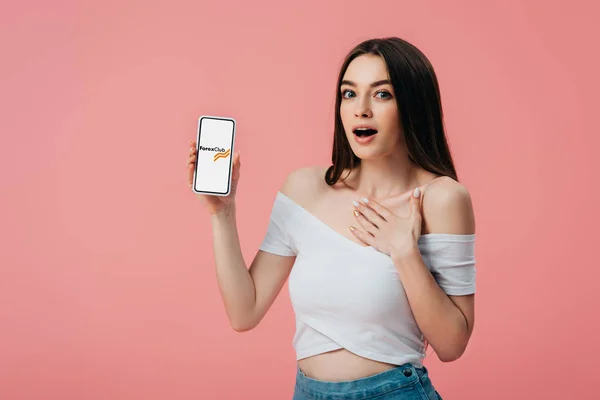 KYIV, UKRAINE - JUNHO 6, 2019: linda garota chocada segurando smartphone com aplicativo de clube forex isolado em rosa — Fotografia de Stock