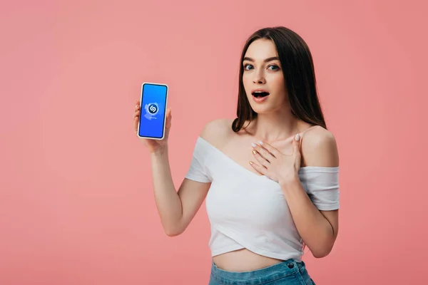 KYIV, UCRANIA - 6 de junio de 2019: hermosa chica sorprendida sosteniendo el teléfono inteligente con la aplicación shazam aislada en rosa — Stock Photo