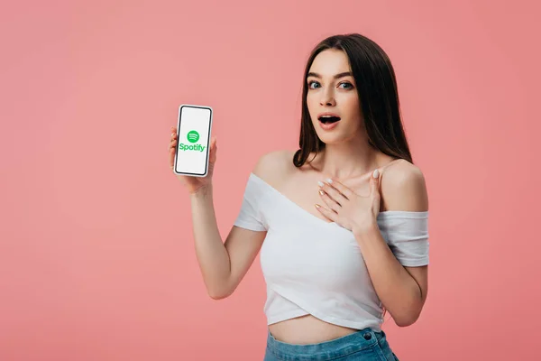 KYIV, UCRANIA - 6 de junio de 2019: hermosa chica sorprendida sosteniendo el teléfono inteligente con la aplicación Spotify aislada en rosa - foto de stock