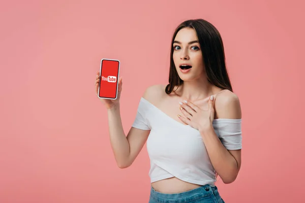 KYIV, UCRANIA - 6 de junio de 2019: hermosa chica sorprendida sosteniendo teléfono inteligente con aplicación de youtube aislada en rosa - foto de stock