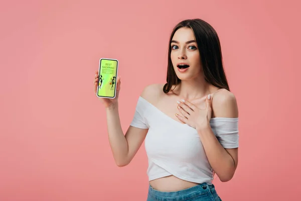 Hermosa chica sorprendida sosteniendo teléfono inteligente con aplicación de compras en línea aislado en rosa - foto de stock