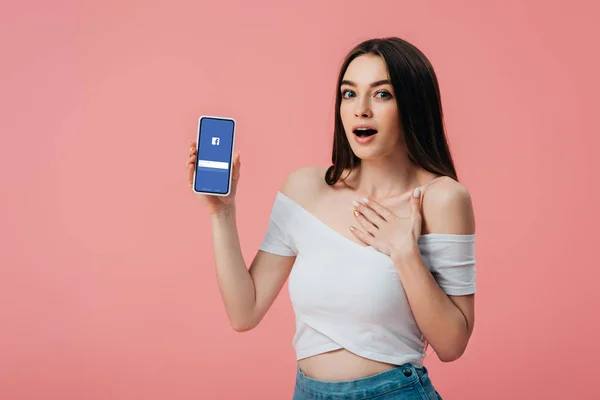 KYIV, UCRANIA - 6 de junio de 2019: hermosa chica sorprendida sosteniendo teléfono inteligente con aplicación de Facebook aislada en rosa - foto de stock