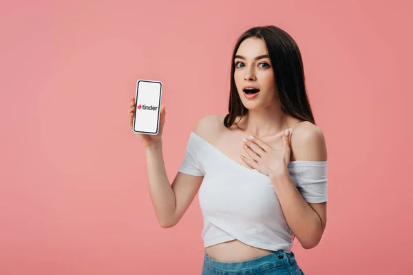 KYIV, UKRAINE - JUNHO 6, 2019: linda garota chocada segurando smartphone com aplicativo Tinder isolado em rosa — Fotografia de Stock