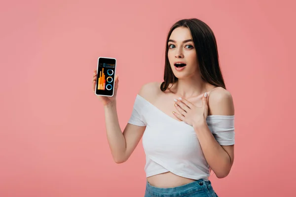 Belle fille choquée tenant smartphone avec application d'analyse financière isolé sur rose — Photo de stock