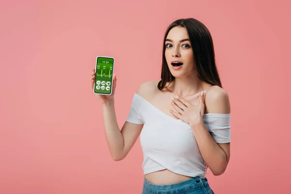 Bela menina chocada segurando smartphone com aplicativo rastreador de saúde isolado em rosa — Fotografia de Stock