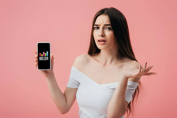 KYIV, UCRANIA - 6 de junio de 2019: hermosa chica confundida sosteniendo el teléfono inteligente con la aplicación Deezer y mostrando un gesto de encogimiento aislado en rosa - foto de stock