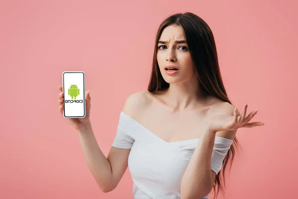 KYIV, UCRANIA - 6 de junio de 2019: hermosa chica confundida sosteniendo el teléfono inteligente con el logotipo de Android y mostrando el gesto de encogimiento aislado en rosa - foto de stock