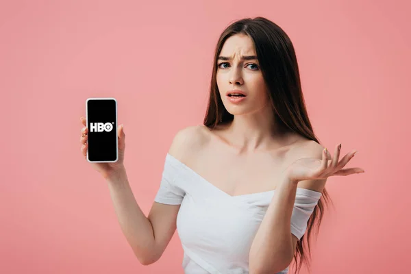 КИЕВ, УКРАИНА - 6 ИЮНЯ 2019 года: красивая смущенная девушка держит смартфон с приложением HBO и показывает жест пожатия, изолированный на розовом — стоковое фото