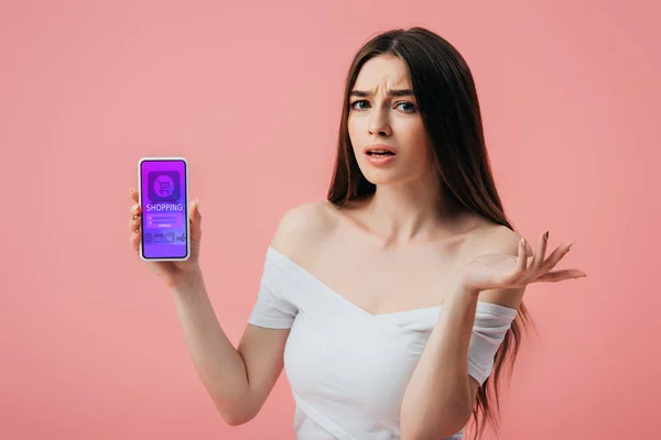 Hermosa chica confundida sosteniendo teléfono inteligente con aplicación de compras en línea y mostrando gesto de encogimiento aislado en rosa - foto de stock
