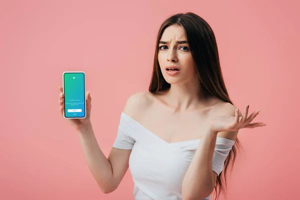 KYIV, UKRAINE - 6 JUIN 2019 : belle fille confus tenant smartphone avec application twitter et montrant geste haussant les épaules isolé sur rose — Photo de stock