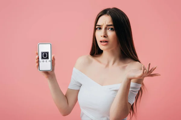 KYIV, UCRANIA - 6 de junio de 2019: hermosa chica confundida sosteniendo el teléfono inteligente con la aplicación Uber y mostrando un gesto de encogimiento aislado en rosa - foto de stock