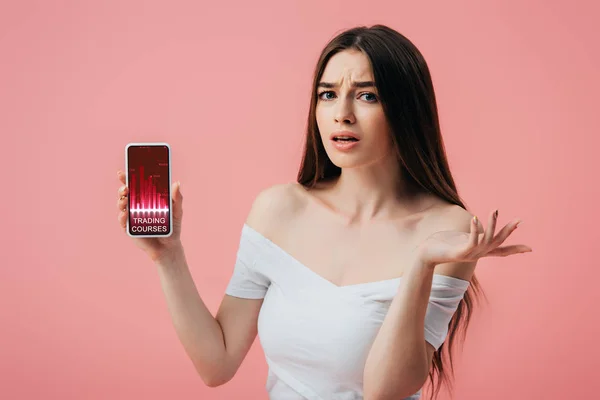 Hermosa chica confundida sosteniendo smartphone con aplicación cursos de comercio y mostrando gesto encogimiento aislado en rosa - foto de stock