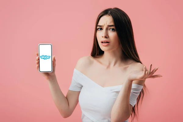 KYIV, UCRANIA - 6 de junio de 2019: hermosa chica confundida sosteniendo el teléfono inteligente con la aplicación Skype y mostrando un gesto de encogimiento aislado en rosa - foto de stock