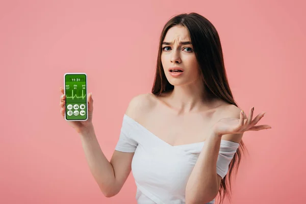 Beautiful confused girl holding smartphone with healthy lifestyle app and showing shrug gesture isolated on pink — Stock Photo