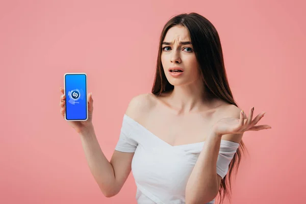 KYIV, UKRAINE - 6 JUIN 2019 : belle fille confus tenant smartphone avec application Shazam et montrant geste haussant les épaules isolé sur rose — Photo de stock