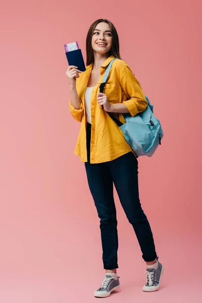 Full length view of beautiful smiling girl with backpack holding passport with air ticket isolated on pink — Stock Photo