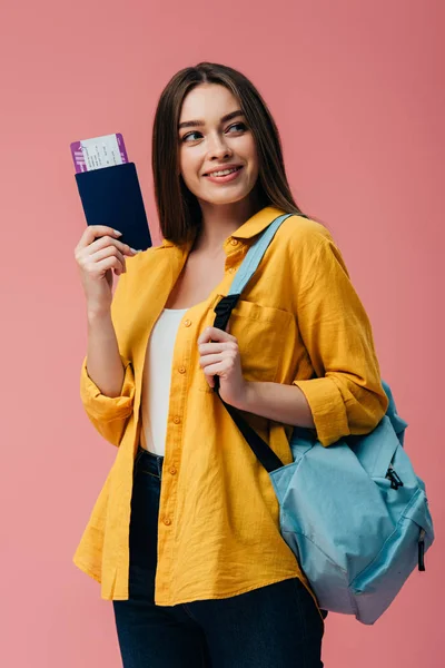 Красива усміхнена дівчина з рюкзаком тримає паспорт з авіаквитком і дивиться ізольовано на рожевий — стокове фото