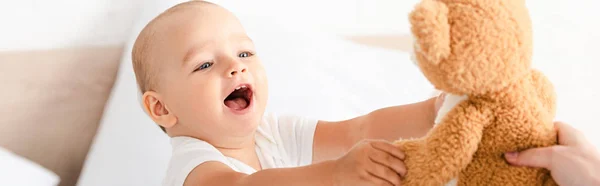 Foto panorámica de lindo niño pequeño sonriendo y levantando las manos para osito de peluche - foto de stock