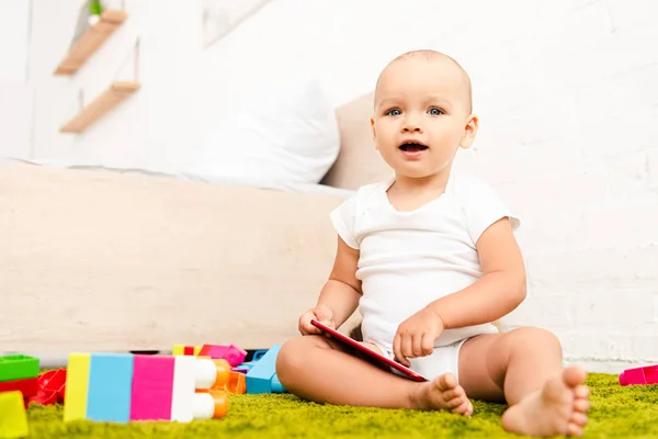 Niedliches Kind mit geöffnetem Mund sitzt auf grünem Boden mit Konstruktion und hält digitales Gerät — Stockfoto