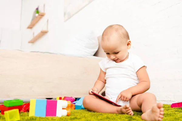 Симпатичный малыш, сидящий и указывающий пальцем на цифровое устройство рядом со строительством на зеленом полу — стоковое фото