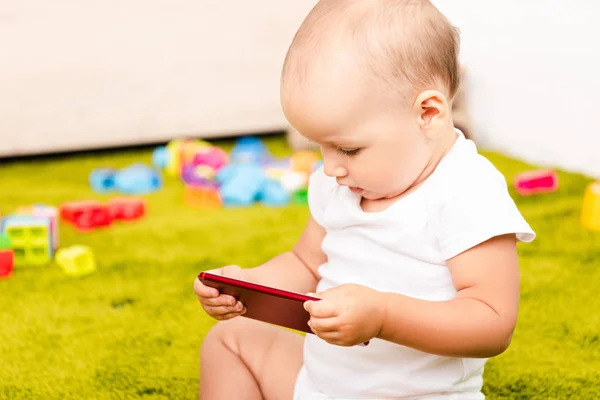 Criança bonito sentado no chão verde com brinquedos e segurando dispositivo digital — Fotografia de Stock