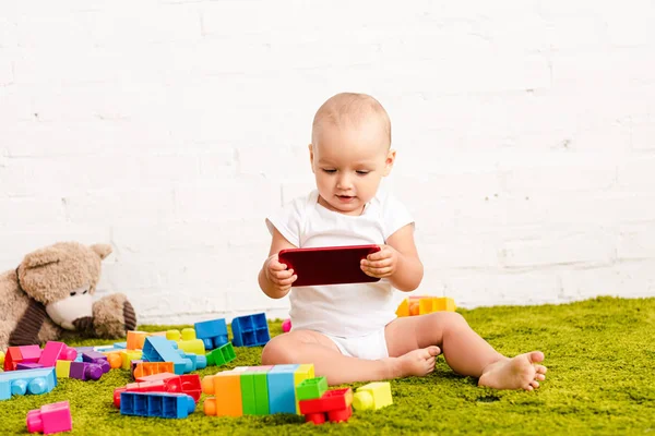 Детеныш, сидящий среди игрушек на зеленом полу и держащий в руках цифровое устройство — стоковое фото
