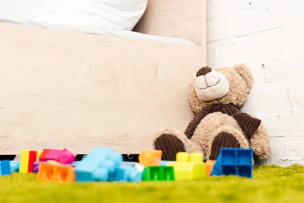 Peluche oso y piezas dispersas de construcción en alfombra verde cerca de la cama de madera - foto de stock