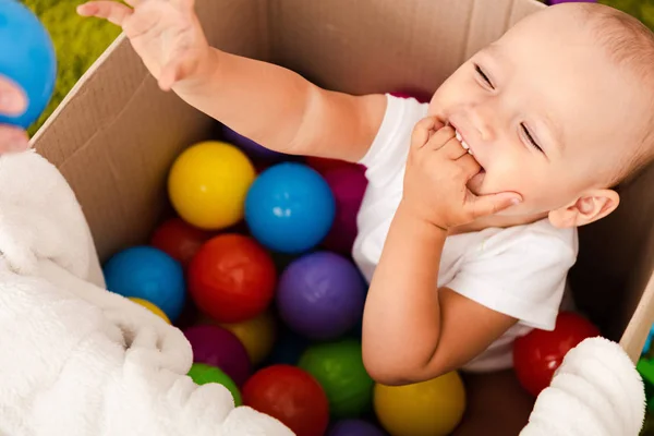 Cute child sitting in cardboard box with colorful balls, laughing and raising his hand up — Stock Photo