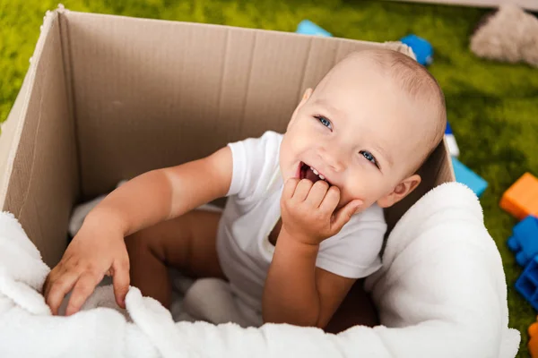 Mignon enfant aux yeux bleus assis et souriant dans une boîte en carton avec couverture — Photo de stock