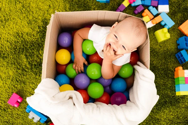 Happy little child sitting in cardboard box with colorful balls and laughing — Stock Photo
