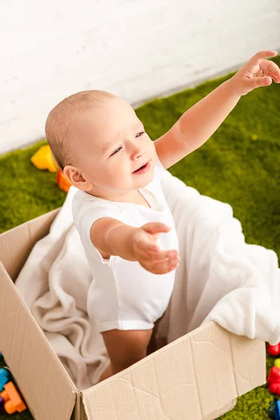 Маленький ребенок стоит в картонной коробке с белым одеялом и поднимая руки — стоковое фото