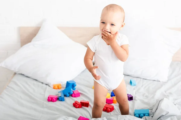 Smiled barefoot baby in white clothes standing on bed with toys and taking fingers into his mouth — Stock Photo