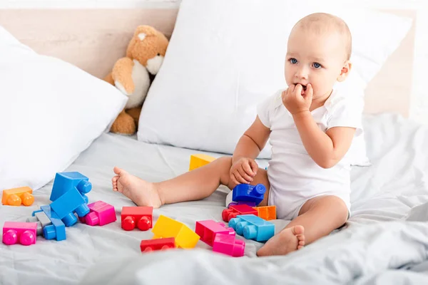 Bebê descalço em roupas brancas sentado na cama com brinquedos e levando os dedos em sua boca — Fotografia de Stock