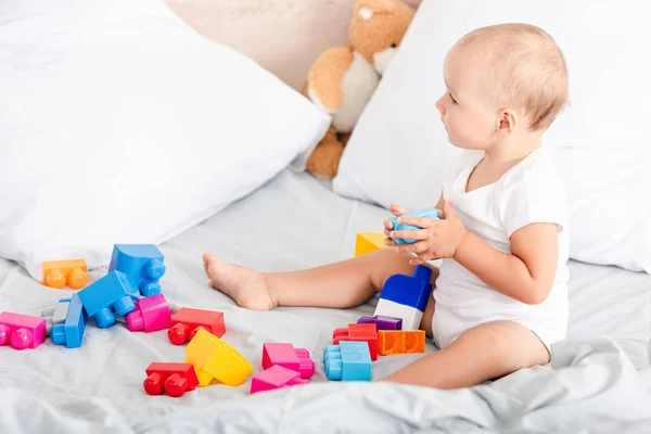 Descalço criança em roupas brancas sentado na cama com travesseiros perto de brinquedos coloridos — Fotografia de Stock