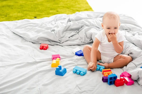 Engraçado bebê descalço em roupas brancas sentado na cama perto de construção colorida e levando os dedos em sua boca — Fotografia de Stock