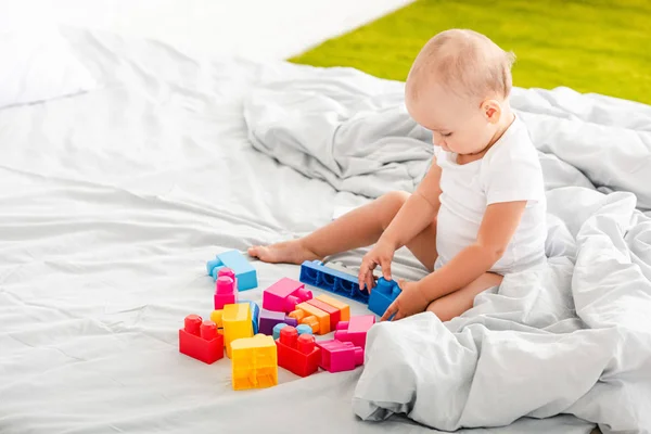 Niedliches Barfußbaby in weißer Kleidung sitzt auf dem Bett und spielt mit buntem Spielzeug — Stockfoto