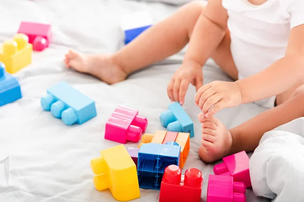 Partial view of barefoot baby in white clothes sitting on bed and playing with bright construction — Stock Photo