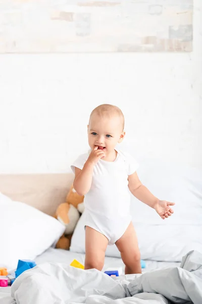 Bebê descalço bonito em roupas brancas em pé na cama com brinquedos e tomando a mão na boca — Fotografia de Stock