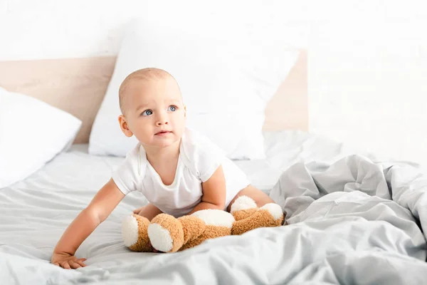Criança bonito em roupas brancas segurando seu urso de pelúcia na cama com travesseiros — Fotografia de Stock