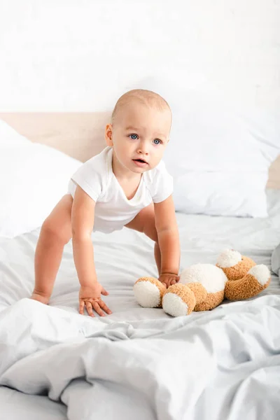 Lindo niño con ropa blanca arrancando su osito de peluche de la cama — Stock Photo