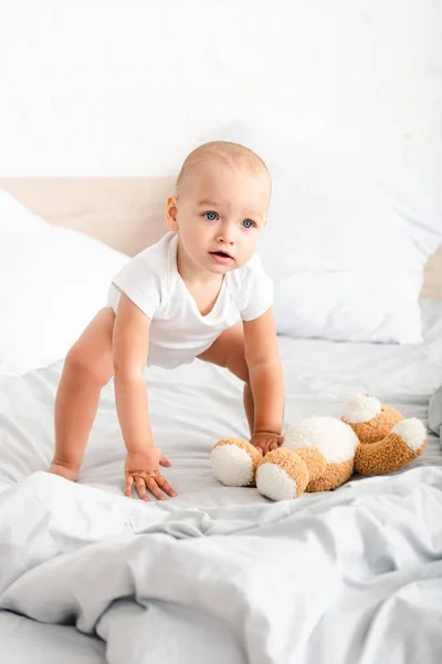 Lindo niño con ropa blanca arrancando su osito de peluche de la cama - foto de stock